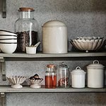 Beige skål INGRID från Strömshaga pryder en rustik hylla, omgiven av andra köksdetaljer. Perfekt för att skapa en varm och hemtrevlig känsla i ditt kök.