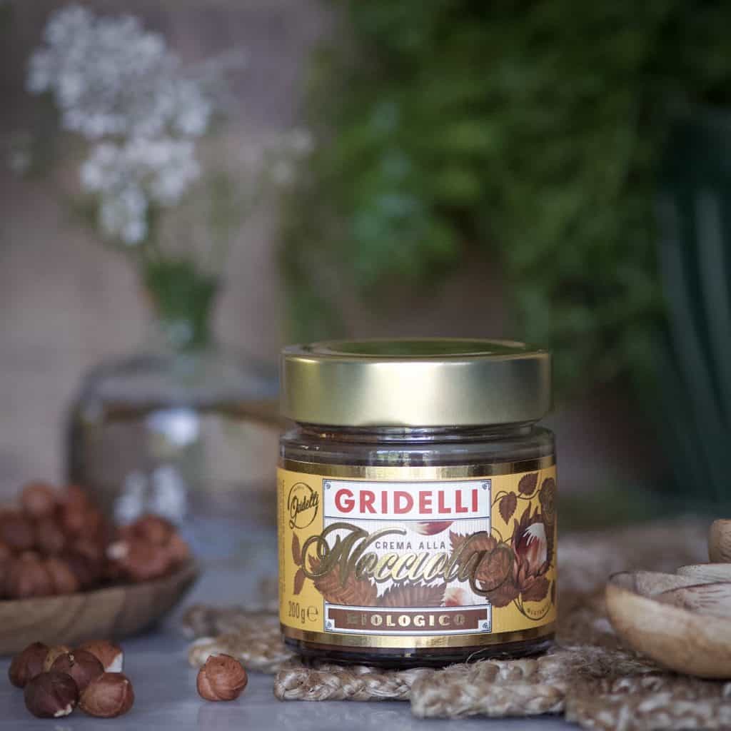 En burk Gridelli Crema alla Nocciola ekologisk hasselnötscreme, omgiven av hasselnötter och rustika dekorativa element.