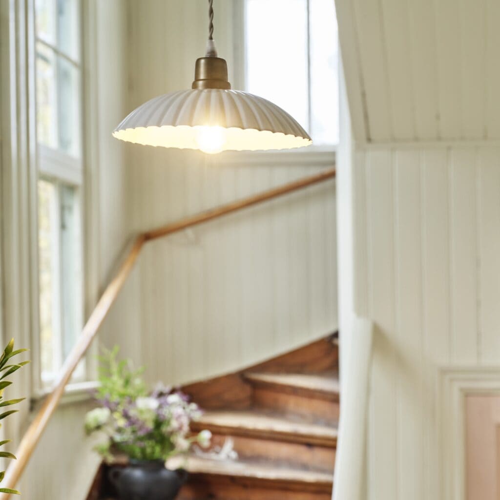 Taklampan INGRID i antik vit från varumärket Strömshaga hänger i en ljus och charmig trappuppgång med trädetaljer och blommor i en vas på ett trappsteg.