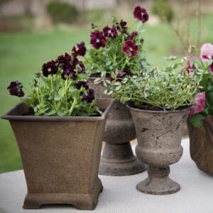 En vacker utomhusmiljö med flera blomkrukor, inklusive den fyrkantiga krukan till vänster som heter EMBLA och kommer från Wikholm form. Krukan är rostfärgad och fylld med frodiga gröna växter och mörklila blommor. I bakgrunden syns fler blomkrukor och en grönskande trädgård.