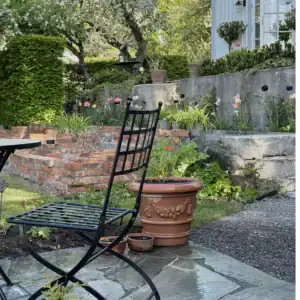 En svart trädgårdsstol vid namn VENICE från varumärket Wikholm form står på en stenlagd uteplats i en lummig trädgård. Bakom stolen syns en terrakottakruka med dekorativa detaljer och en upphöjd rabatt med blommande växter och gröna buskar. I bakgrunden finns ett tegelmurat område och en vit byggnad omgiven av grönska.