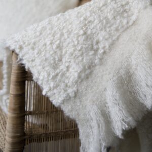 Närbild på pläden NALIA från Wikholm Form i mjuk, vit bouclé. Pläden hänger över en rottingstol och visar tydligt den texturerade ytan och de mjuka fransarna längs kanten.