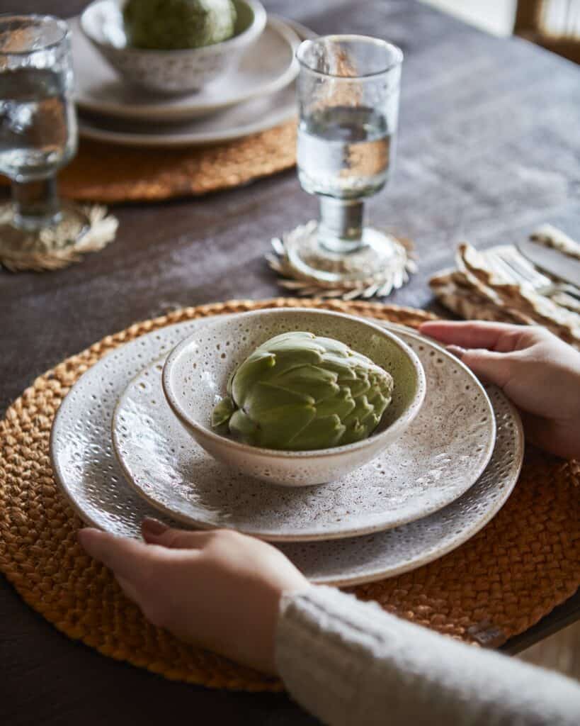 Närbild på ett vackert dukat bord med jordnära keramiktallrikar, vävda underlägg och en färsk kronärtskocka som mittpunkt. Scenen fångar värmen och texturen i en mysig höstdukning, med fokus på naturliga material och inbjudande toner.
