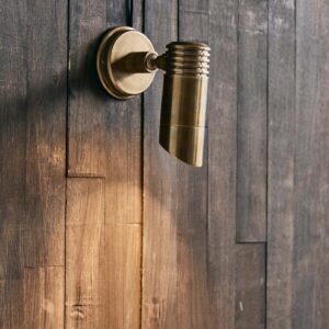 Bragi vägglampa från Nordal i mässing, monterad på en mörk trävägg. Elegant utebelysning med justerbar spot som åldras vackert med patina över tid.