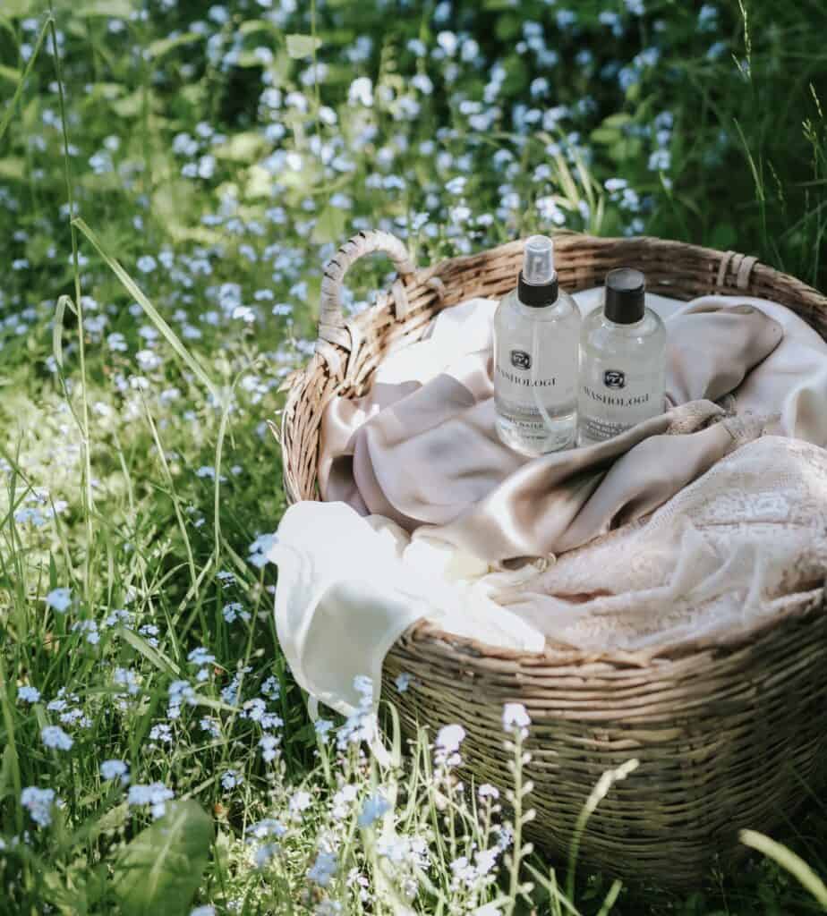 Washologi linnevatten och tvättmedel i en korg på gräset, omgiven av blommande blommor – miljövänliga produkter som ger dina kläder en fräsch och härlig doft.