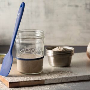 Kilner-glasburk fylld med surdeg, placerad på en skärbräda tillsammans med en blå silikonspatel och mjöl i bakgrunden.