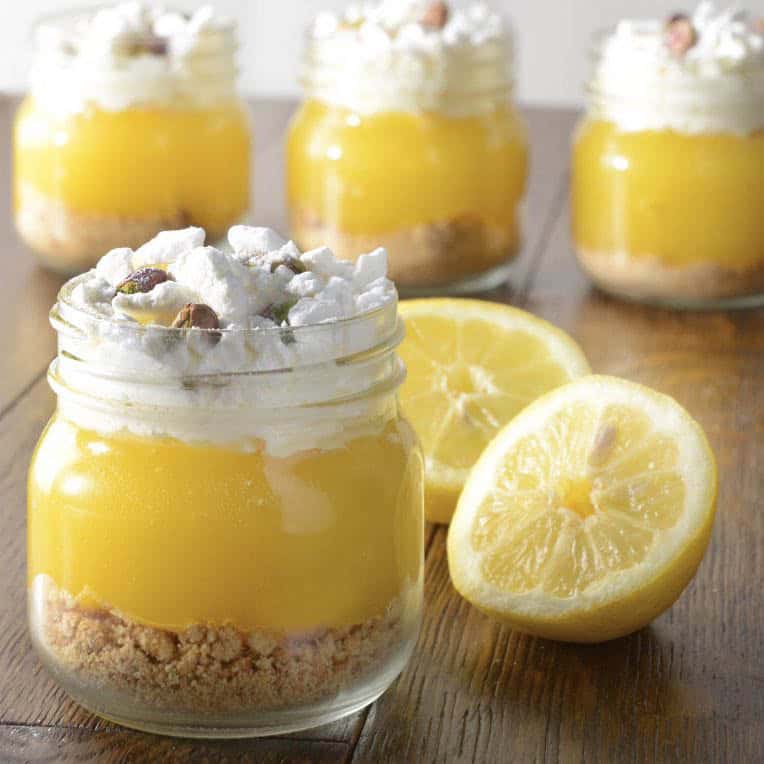 Kilner-burkar fyllda med citronkräm, toppade med grädde, maräng och nötter, bredvid halverade citroner.