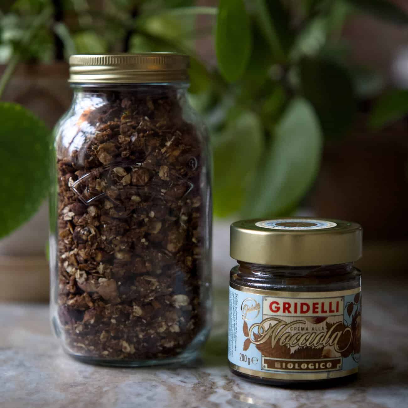 Glasburk fylld med choklad- och hasselnötsgranola bredvid en burk Gridelli hasselnötskräm, placerade på ett marmorbord med gröna växter i bakgrunden.