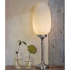 Bordslampa Evald från Strömshaga med avlång glaskupa i frostat vitt och en dekorativ fot i förnicklad metall, placerad mot en rustik bakgrund med glasvaser och gröna växter.