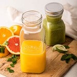 Kilner glasflaskor fyllda med färskpressad juice och grön smoothie, omgivna av citrusfrukter och lime på en rustik träbricka.