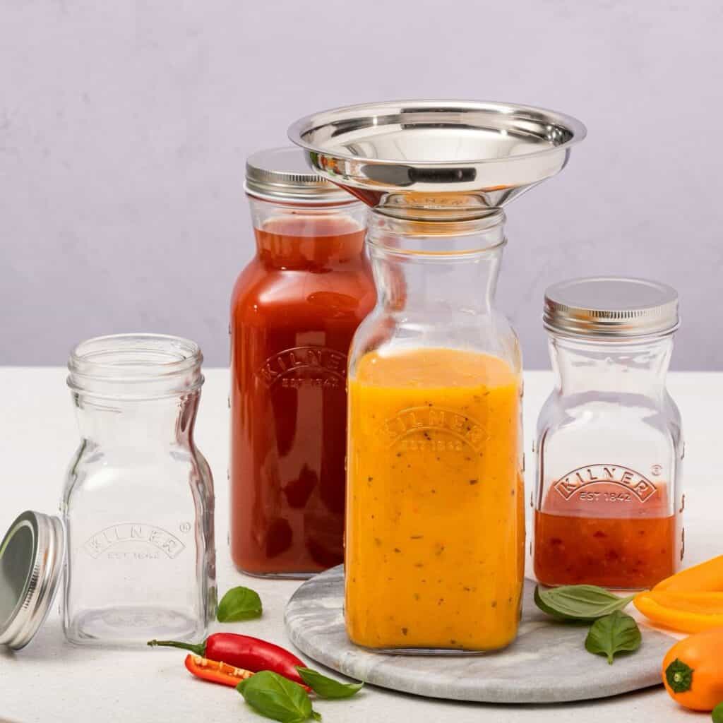Kilner glasflaskor i olika storlekar, fyllda med såser och drycker, tillsammans med en praktisk rostfri tratt för enkel påfyllning.