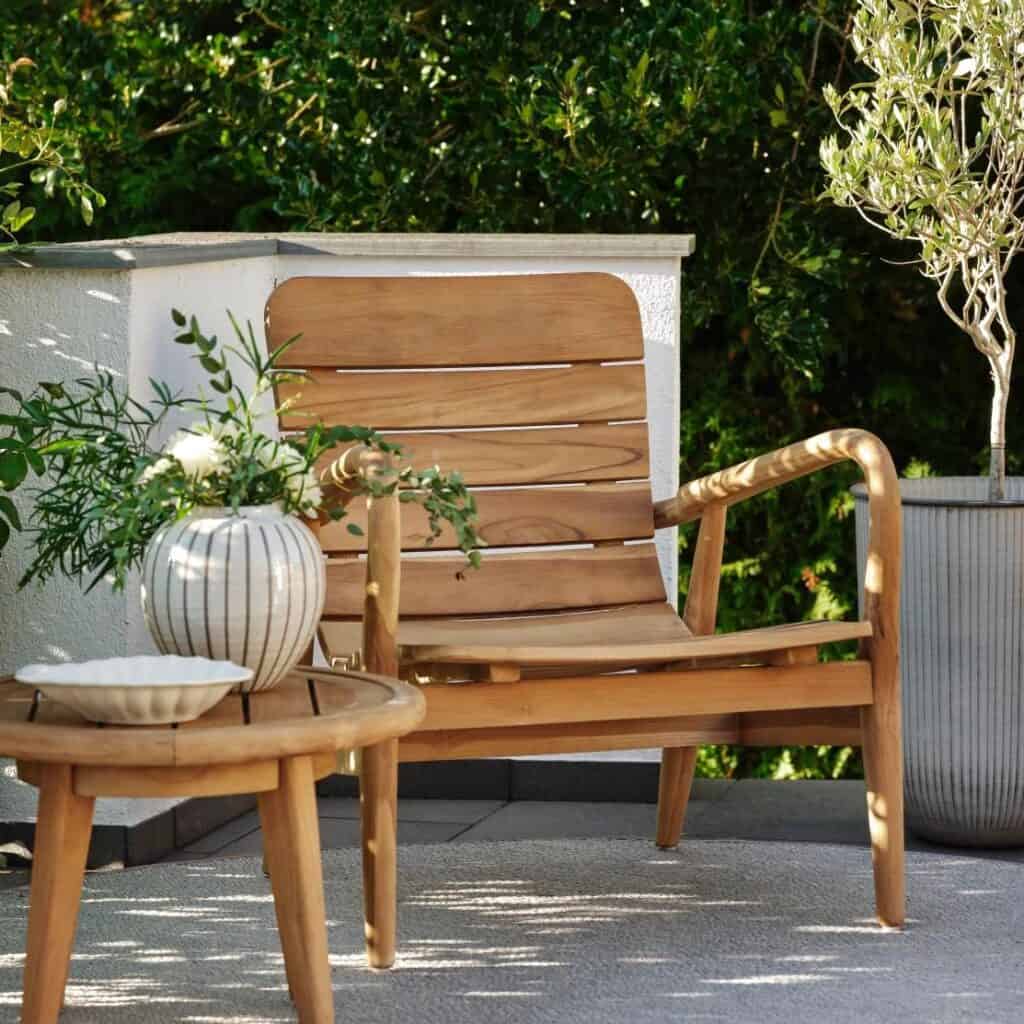 Loungefåtölj i teak placerad på en solig uteplats, omgiven av grönska och dekorerad med ett litet sidobord och blomvas.