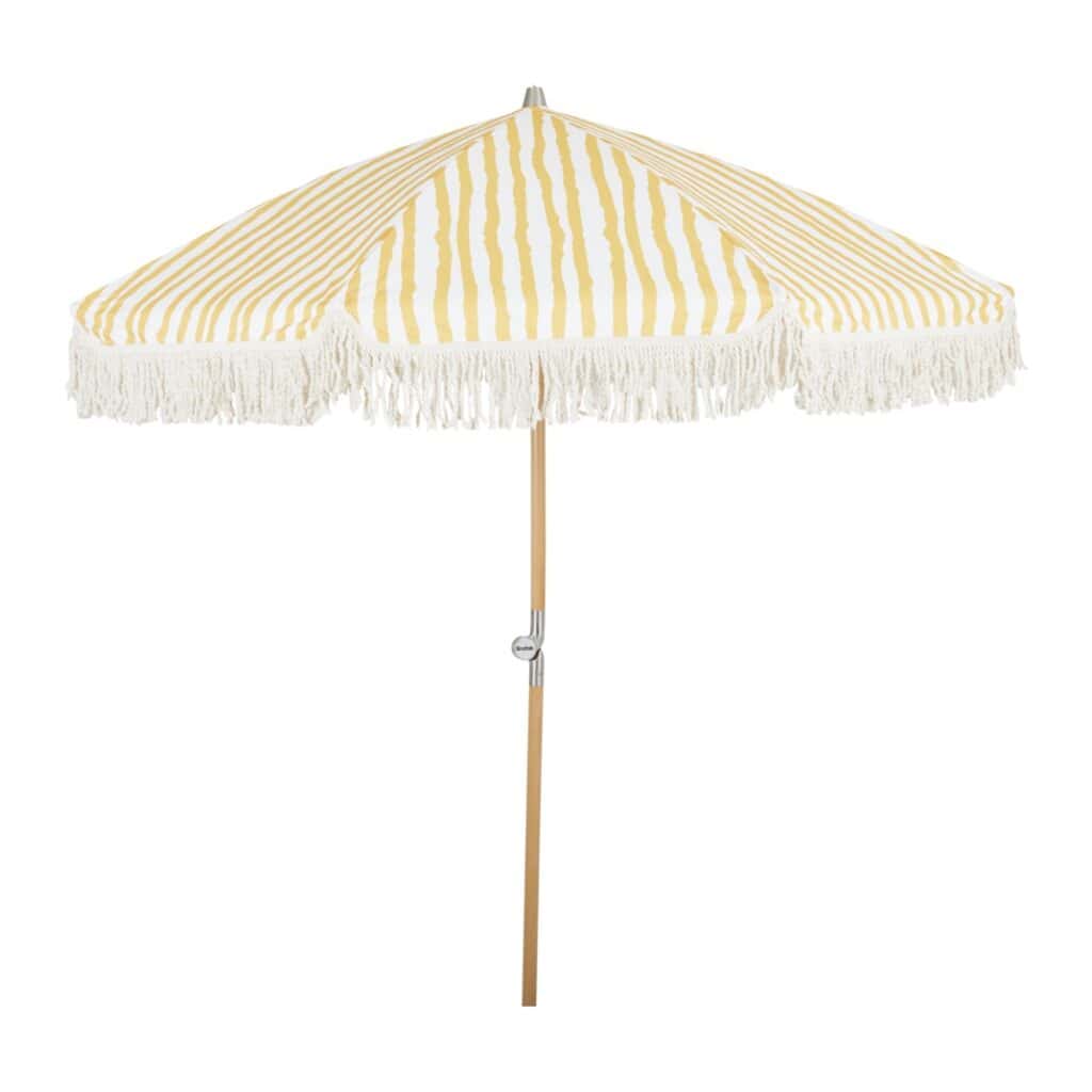 Gulrandigt parasoll Gatsby från Brafab med fransdetaljer och lutningsbar funktion, stående på en naturlig trästång.
