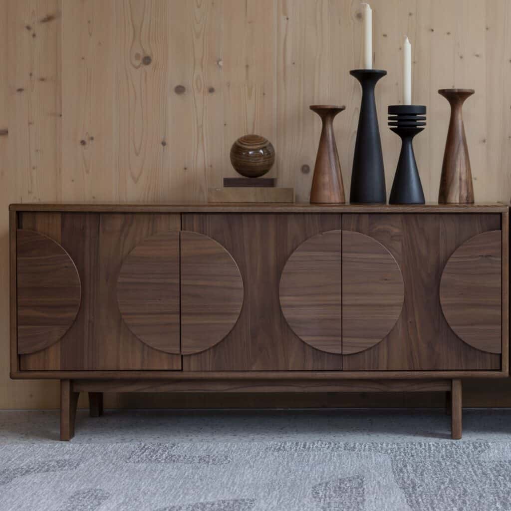 Sidobord Groove i valnötsfanér med tre dörrar och runda detaljer, dekorerat med stilrena vaser och ljusstakar i ett modernt träinrett rum.