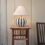 Lampfot ALBA från Strömshaga med svartvit randig glasfot och beige lampskärmen Hedvig, placerad på ett rustikt träbord mot en ljus trävägg.