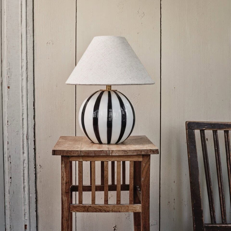 Lampfot ALBA från Strömshaga med svartvit randig glasfot och beige lampskärmen Hedvig, stående på ett rustikt träbord mot en ljus trävägg.