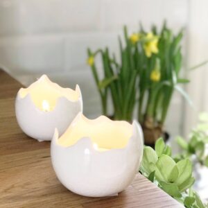 Två vita ljuslyktor i form av spruckna äggskal från Bloomingville med tända ljus, placerade på en träyta med påskliljor och gröna växter i bakgrunden.