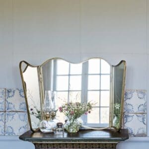 Vikbar spegel i antik mässing från Strömshaga, placerad på ett gammaldags bord med dekorativa blommor och oljelampa framför. Spegeln reflekterar ett fönster som skapar ett ljust och luftigt intryck.