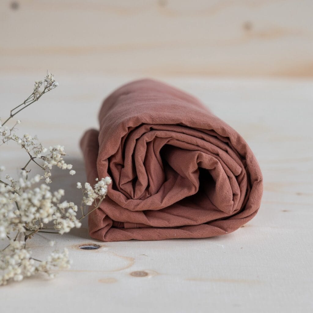 Underlakan ekologisk bomull Terracotta 240×260 cm – hoprullat på träbord med vita blommor i förgrunden