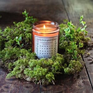 Doftljus i glas med kalenderetikett från Strömshaga, tänd låga och mjukt ljus i julig miljö med mossa och naturdetaljer.