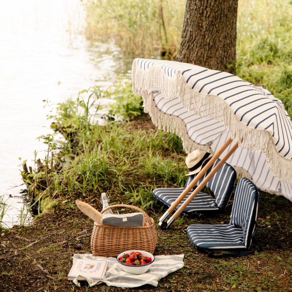 Randigt parasoll AUGUST i offwhite och blått vid sjö med picknick och sittdynor