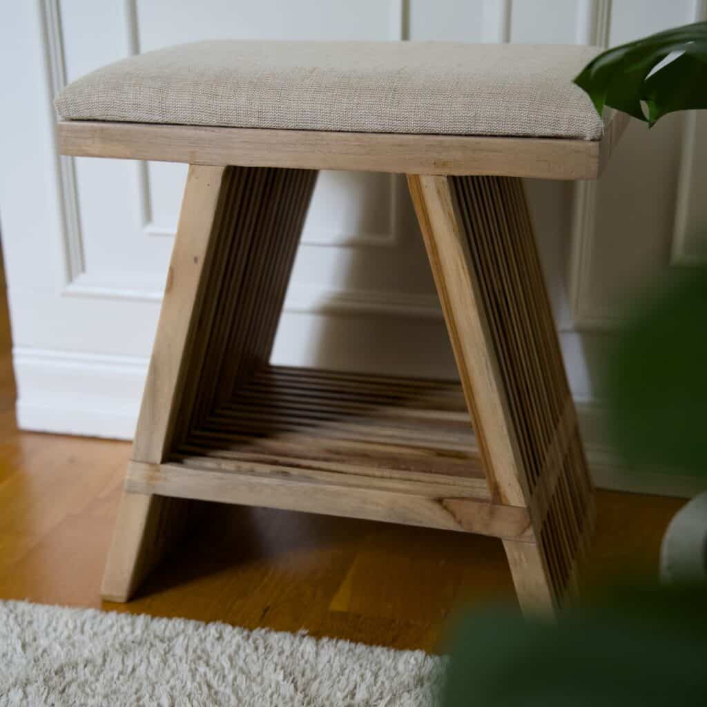 Pall i teak med beige tygsits som visar ribbad träkonstruktion, varm träton och placering i ljus hemmiljö.