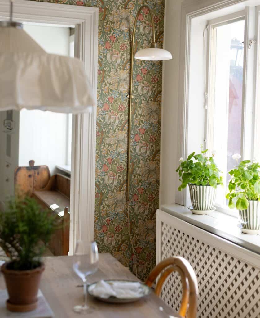 Lantligt rum med pelargoner i fönstret, bordslampa, träbord och blommig tapet
