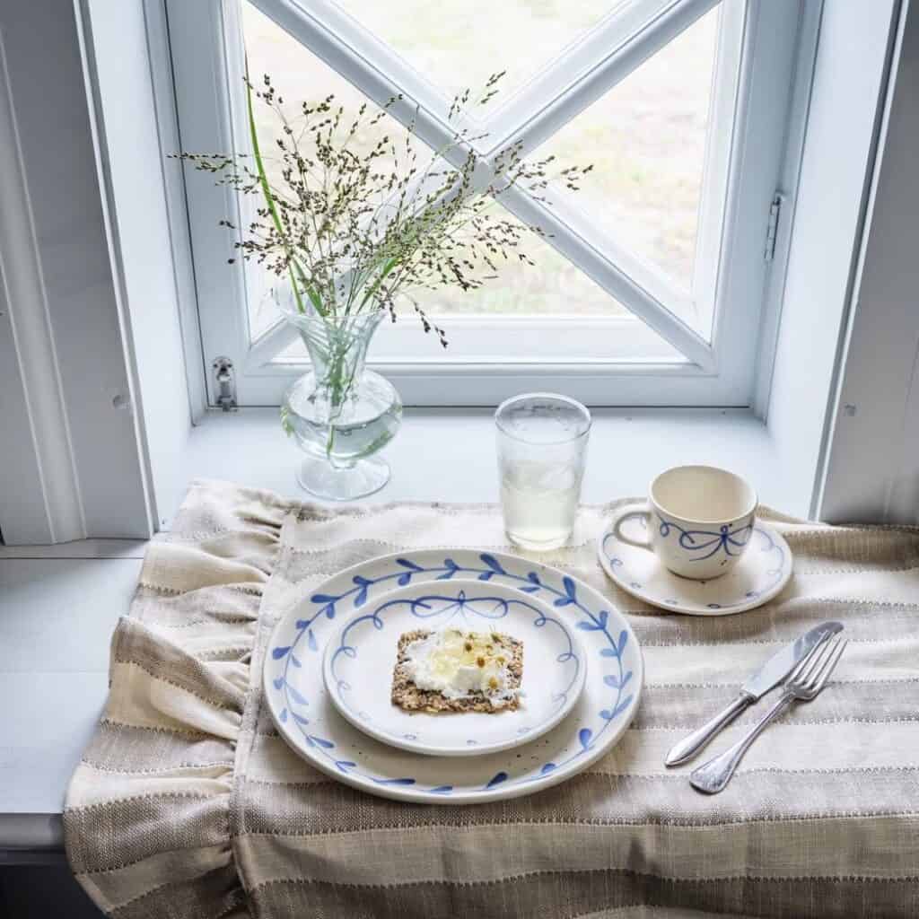 Glasvas Annie i klart glas med gröna strån vid en dukning i fönster.