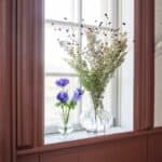 Glasvas Annie i klart glas med lila blommor i ett fönster.