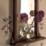 Glasvas Annie i klart glas med lila blommor framför en brun spegel.