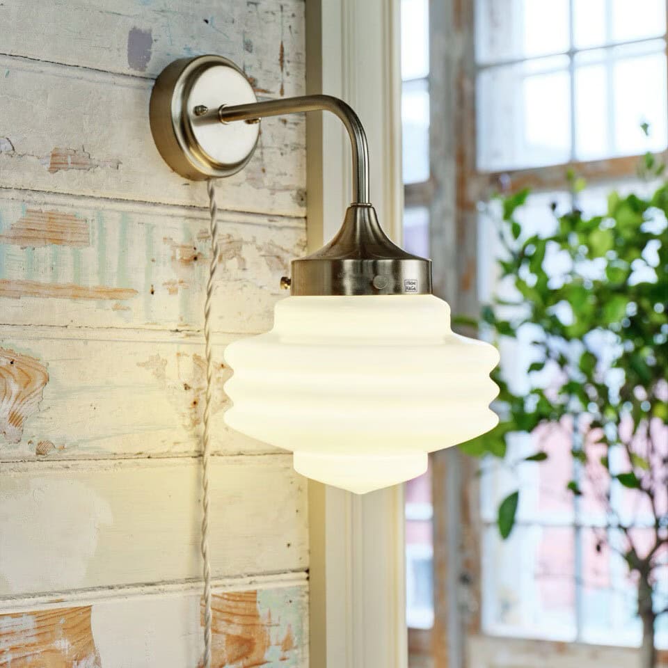 Vägglampa Valter i mässing med vitt opalglas tänd på vägg i ljus inomhusmiljö